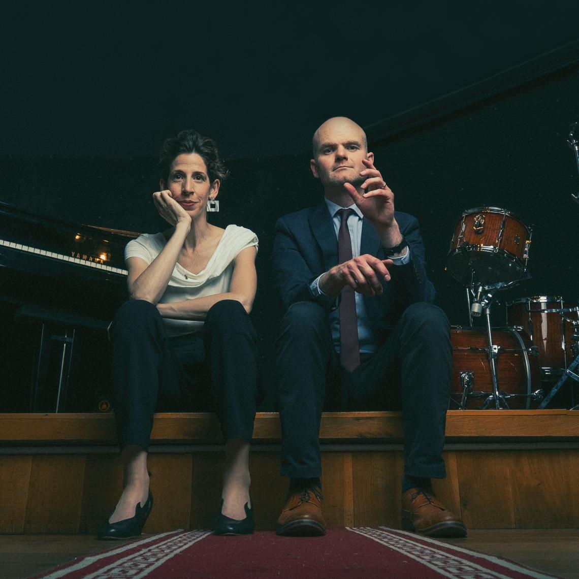 Eine Frau und ein Mann sitzen am Rand einer Bühne vor einem Schlagzeug und einem Klavier