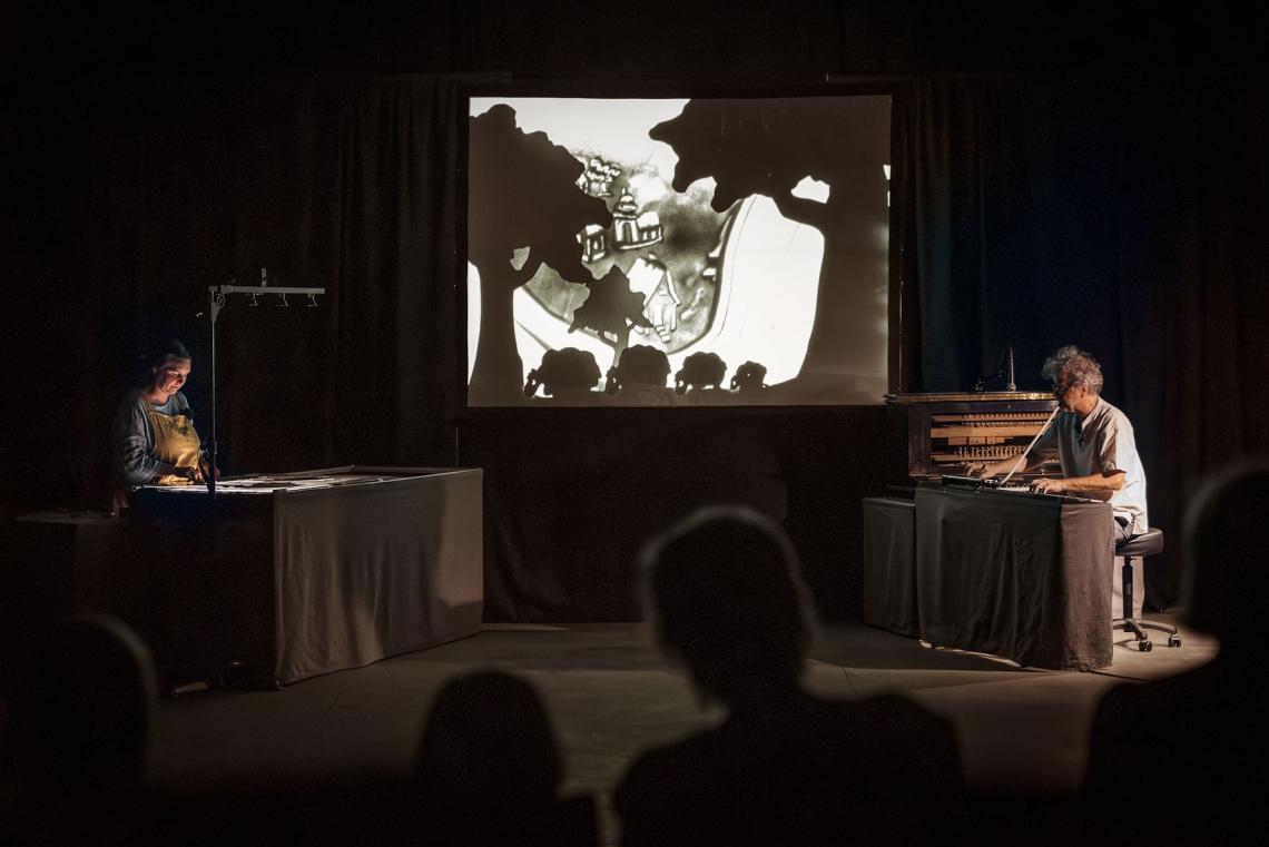 Zwei Puppenspielerinnen auf einer Bühne, deren Hintergrund eine schwarz-weiße Projektion bildet.