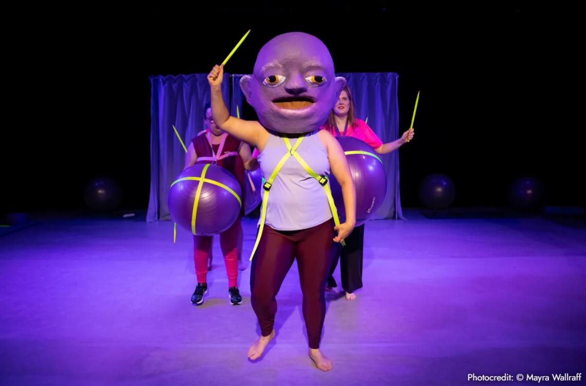 Drei Frauen mit Trommelstöcken stehen auf einer Bühne. Eine hat einen sehr großen Kopf, die anderen haben große schwangere Bäuche.