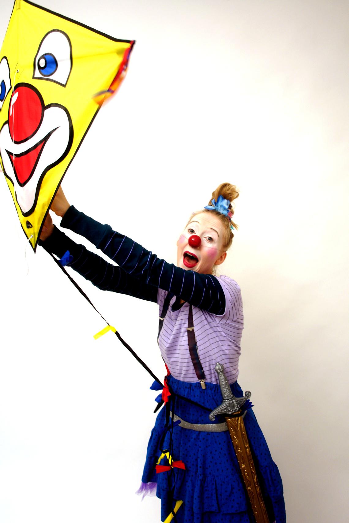 Eine junge Frau mit Clownsgesicht hält einen Drachen fest, auf dem ebenfalls ein Clownsgesicht zu sehen ist.