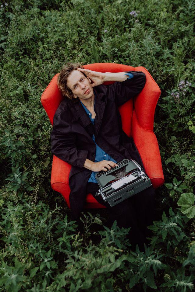 Ein Mann sitzt mit einer Schreibmaschine auf dem Schoß in einem roten Sessel auf einer Wiese.