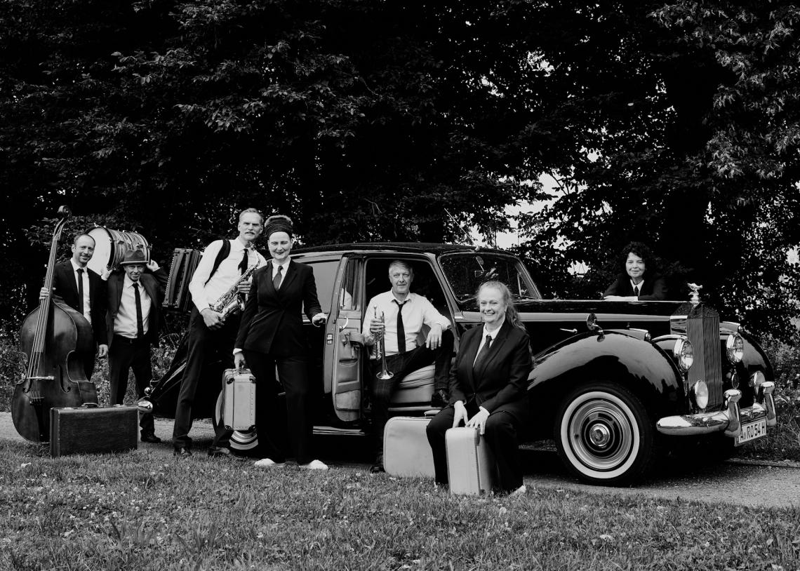 Eine Swingband in amerikanischer Kleidung der 40er-Jahre posiert vor einem Oldtimer.