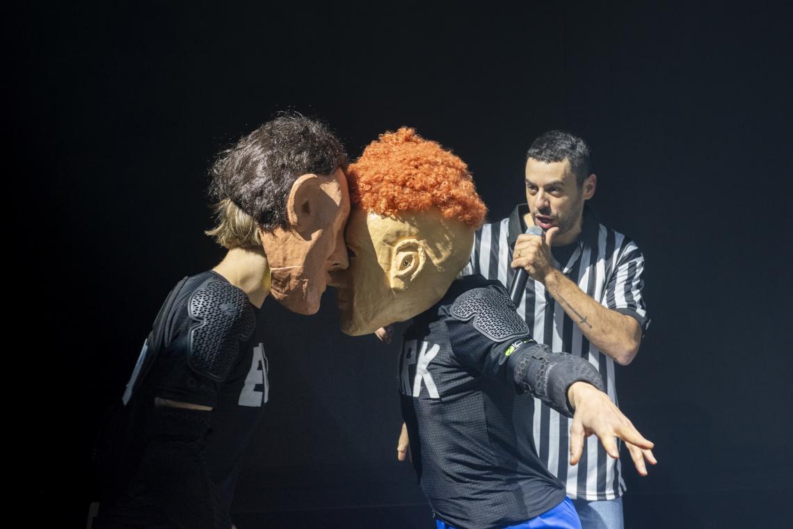 Zwei Figuren mit Masken kämpfen miteinander, während ein Schiedsrichter zusieht.