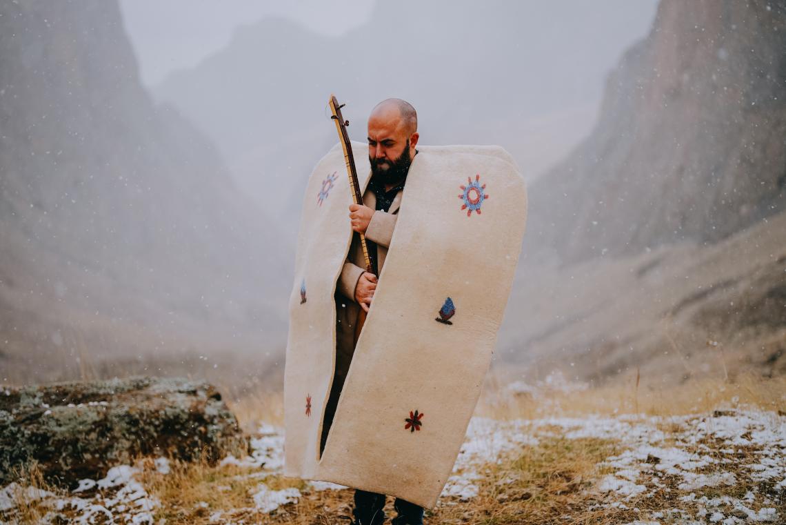 Ein Mann steht in traditioneller anatolischer Kleidung mit einem Saiteninstrument auf einem Berg.