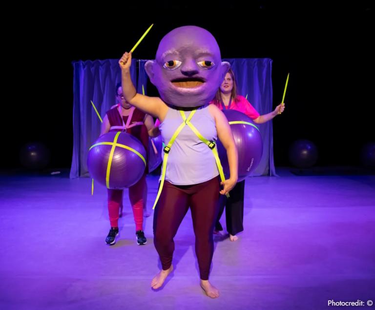 Drei Frauen mit Trommelstöcken stehen auf einer Bühne. Eine hat einen sehr großen Kopf, die anderen haben große schwangere Bäuche.