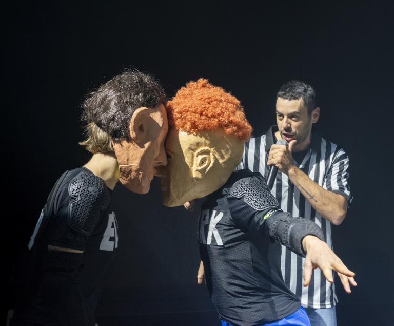 Zwei Figuren mit Masken kämpfen miteinander, während ein Schiedsrichter zusieht.