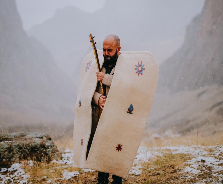 Ein Mann steht in traditioneller anatolischer Kleidung mit einem Saiteninstrument auf einem Berg.
